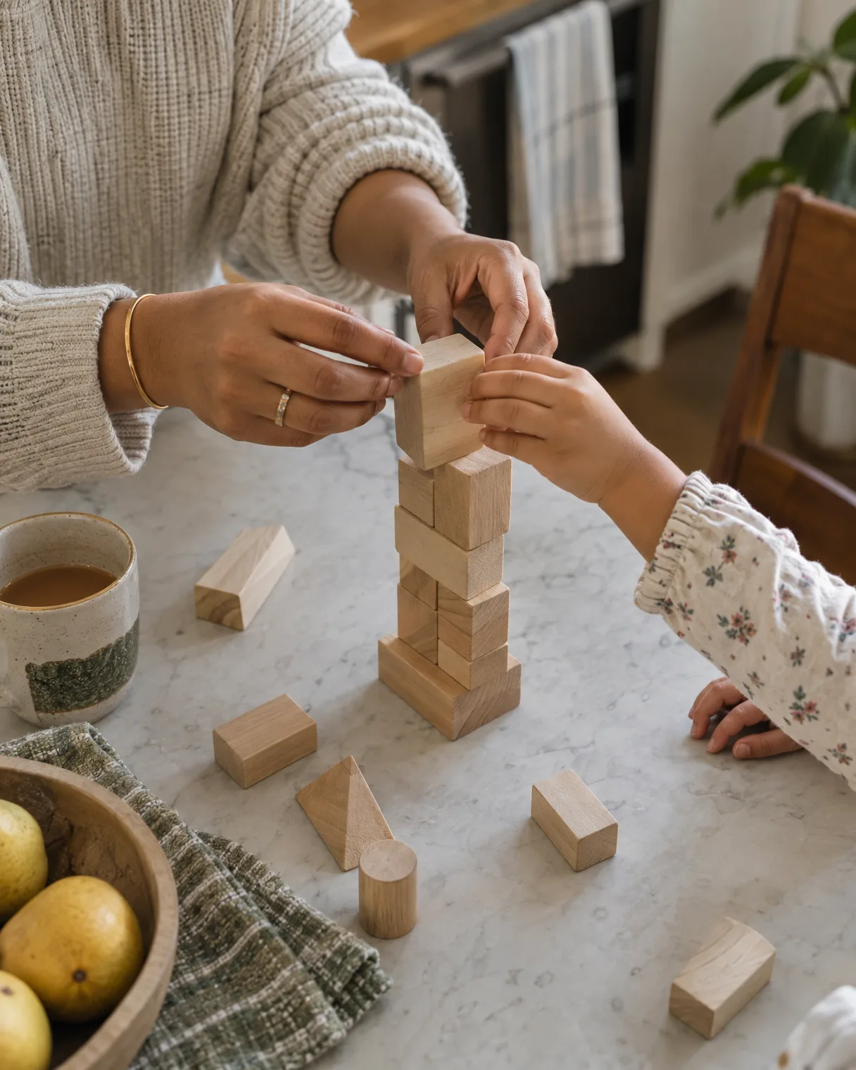 A parent’s hands and a child’s hands building with wooden blocks at a kitchen table